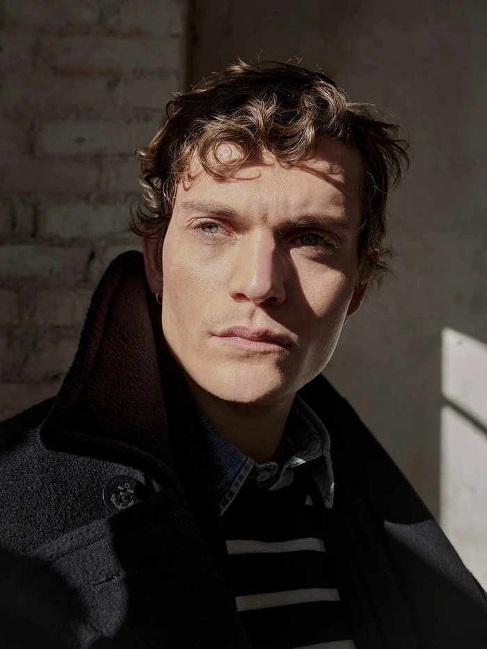 Close-up portrait of a male model wearing a dark coat and striped sweater with strong natural sunlight casting shadows.