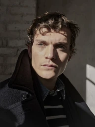 Close-up portrait of a male model wearing a dark coat and striped sweater with strong natural sunlight casting shadows.
