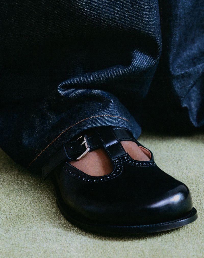 Close-up of a person wearing blue jeans and a black T-strap shoe with a silver buckle, standing on a light green carpet.