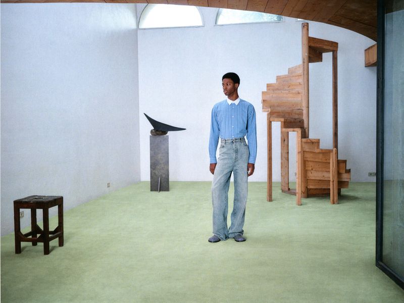 Young Black man in blue striped shirt and light jeans standing in an open, minimalist interior with wood stairs and sculpture.