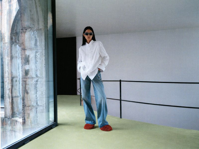A stylish woman in a white button-down shirt and blue jeans stands indoors near a concrete wall and large window for a fashion editorial.