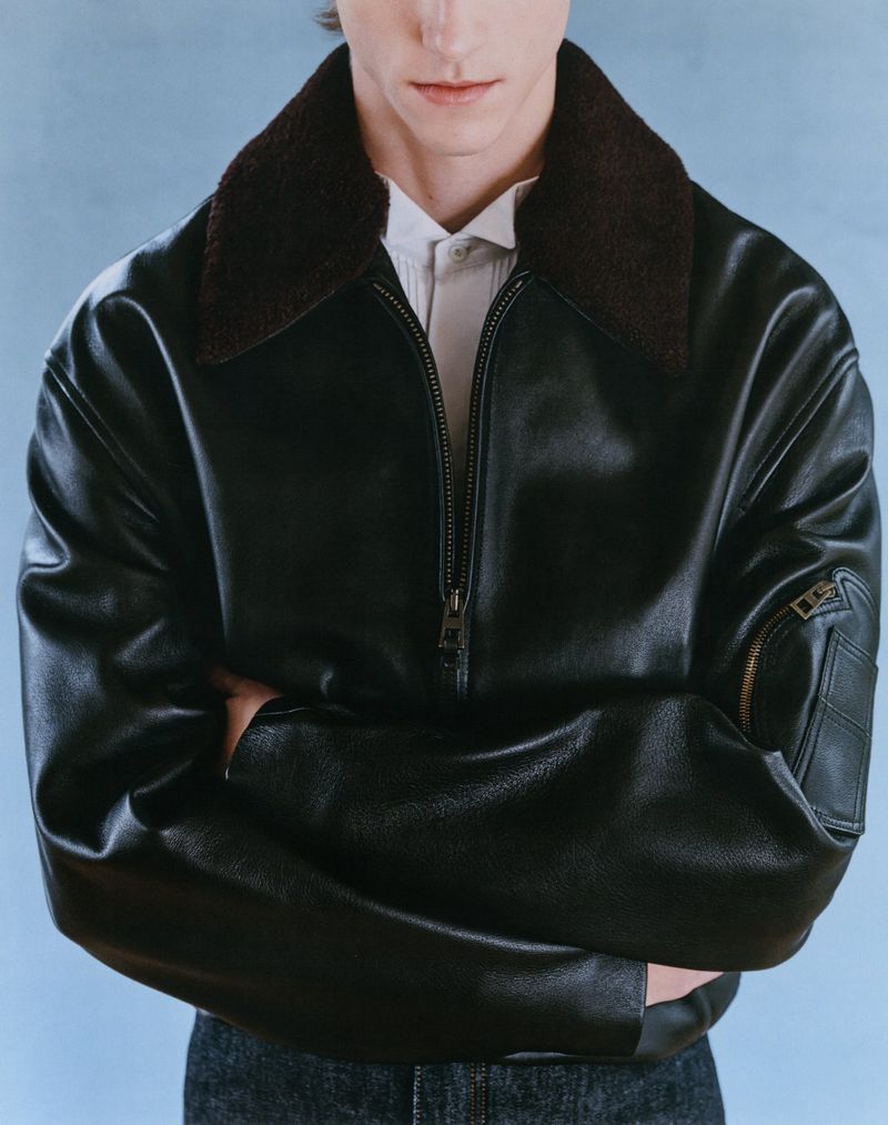 Male model poses in a black leather aviator jacket with a shearling collar and jeans against a light blue backdrop in a studio shot.
