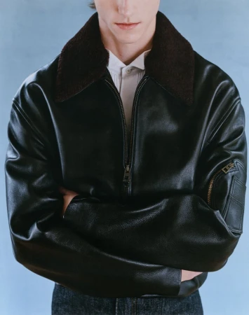 Male model poses in a black leather aviator jacket with a shearling collar and jeans against a light blue backdrop in a studio shot.