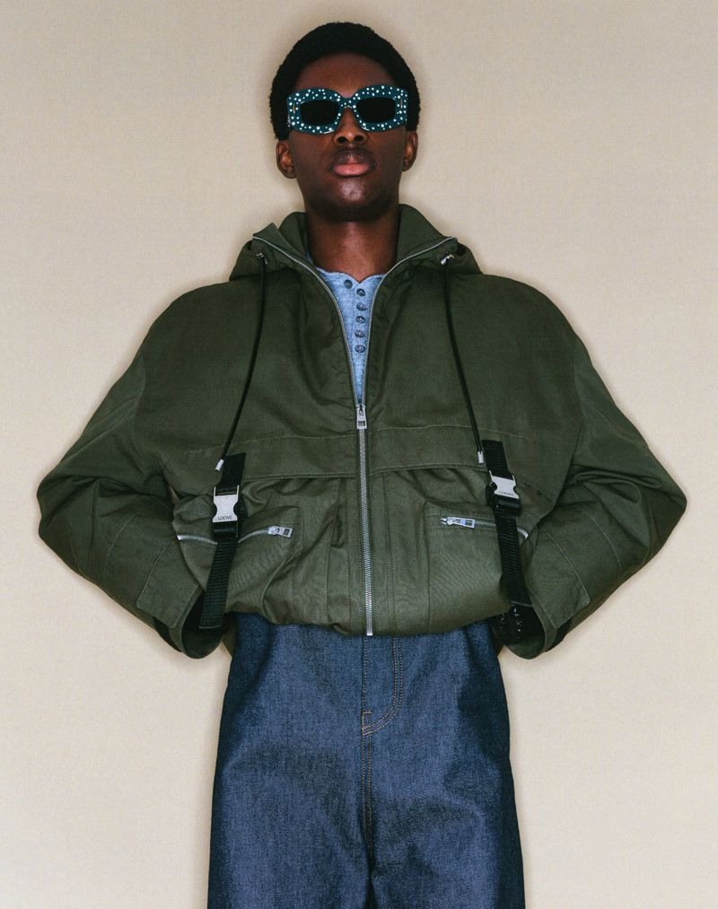 Dark-skinned adult posing indoors wearing a green jacket, blue jeans, and patterned sunglasses against a neutral background.