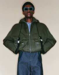 Model wearing an olive green hooded jacket and dark blue denim trousers against a neutral background
