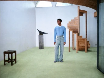 Nonso Ojukwu standing in a room with light green carpet, wearing a blue collared shirt and wide-leg jeans near a wooden spiral staircase.
