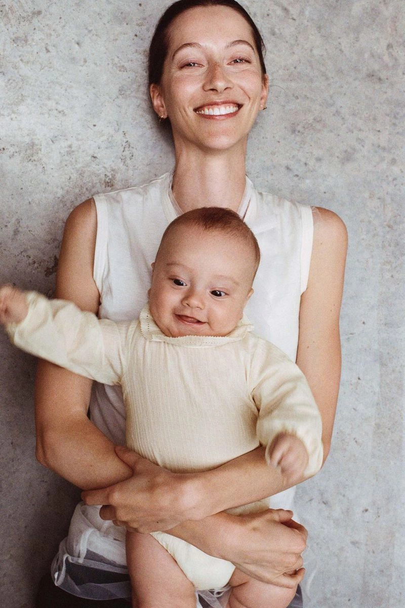 Model Alana Zimmer smiling while holding a baby dressed in a cream long-sleeved onesie in front of a textured concrete wall.