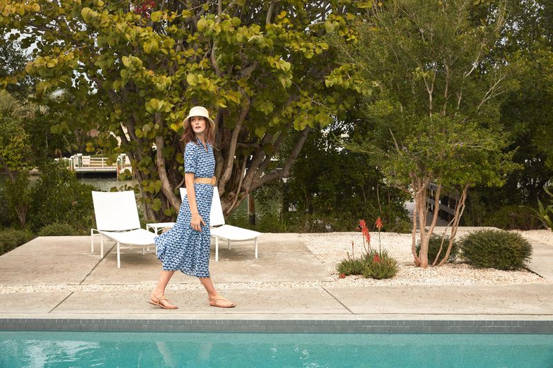 An adult woman in a blue patterned dress and hat walks near a turquoise swimming pool, with lounge chairs and green trees in the background.