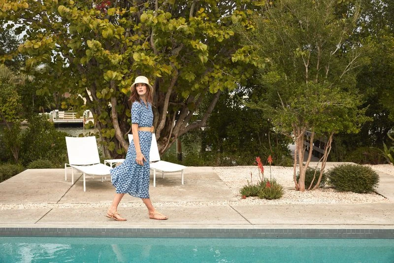 An adult woman in a blue patterned dress and hat walks near a turquoise swimming pool, with lounge chairs and green trees in the background.