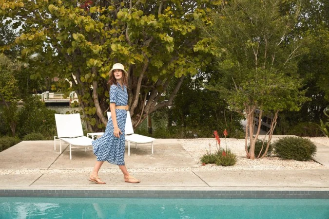 An adult woman in a blue patterned dress and hat walks near a turquoise swimming pool, with lounge chairs and green trees in the background.