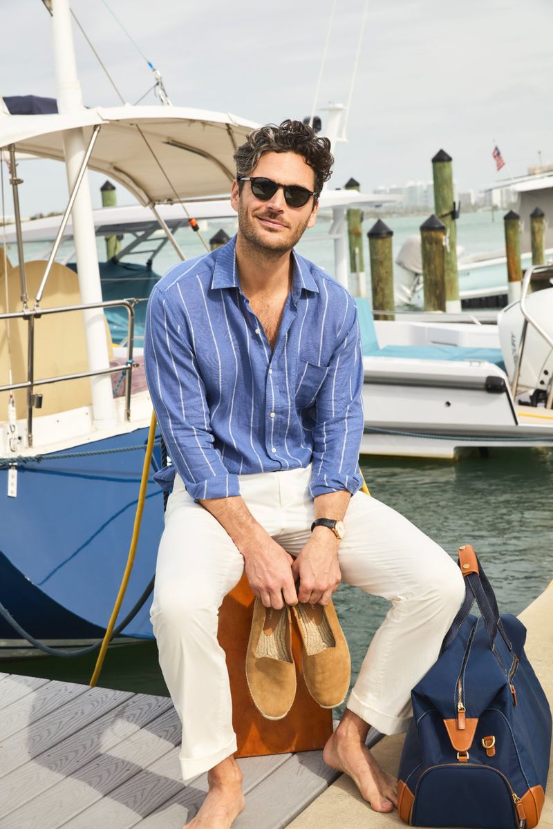 Man sits on an orange crate on a dock in front of a boat wearing a blue striped shirt and holding a pair of tan shoes.