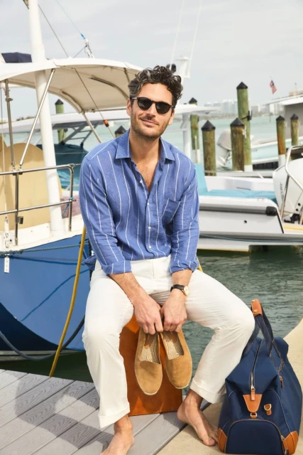 Man sits on an orange crate on a dock in front of a boat wearing a blue striped shirt and holding a pair of tan shoes.