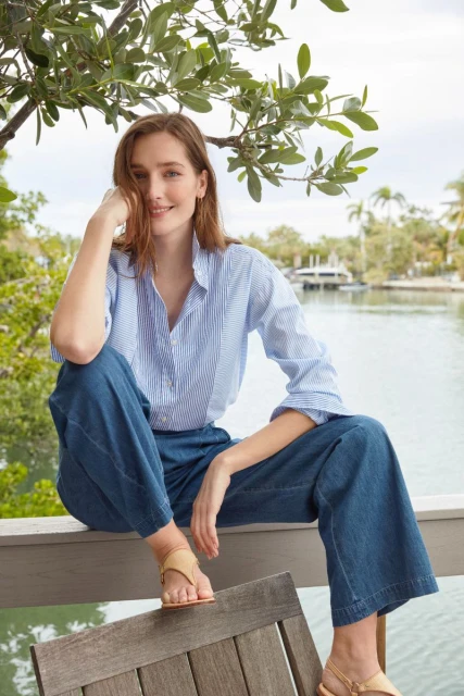 A woman poses in denim and a striped shirt, seated on a wooden structure near a tranquil river on a bright day.