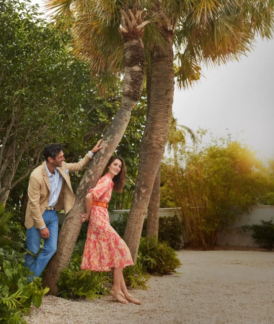 A couple poses beneath palm trees in a sun-drenched outdoor setting; the man in a tan jacket and the woman in a peach floral dress.