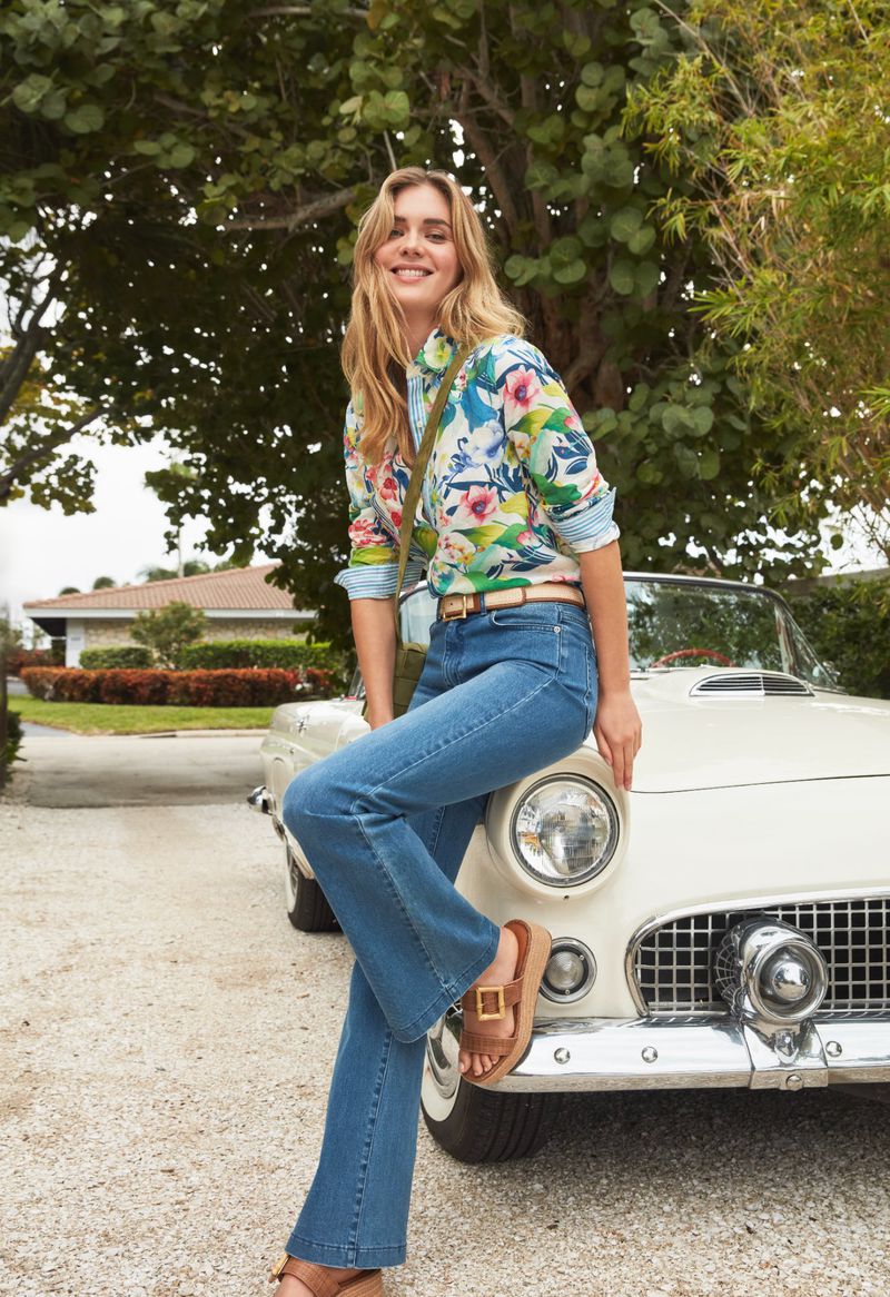 Smiling woman in floral blouse and jeans leans against white vintage car in outdoor setting with leafy trees and residential buildings.