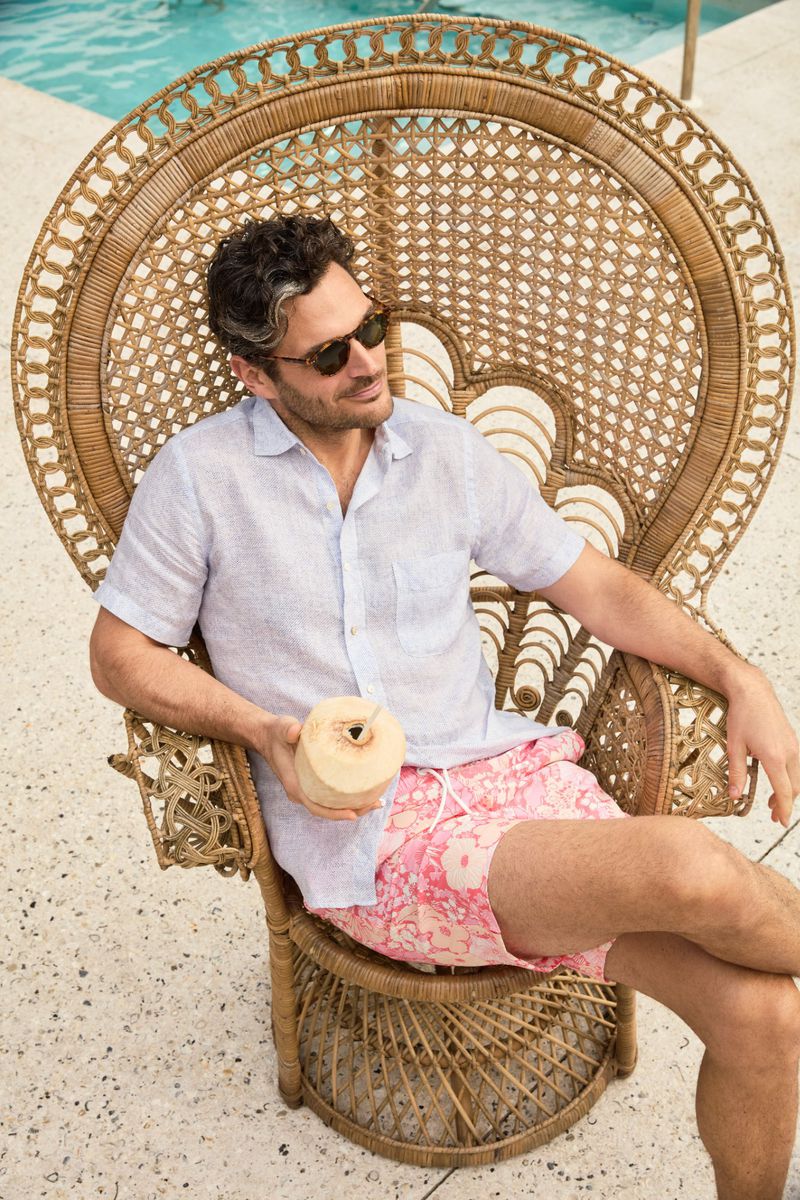 Adult man in a woven chair by a pool, wearing sunglasses and a patterned shirt, holding a coconut drink in a tropical setting.