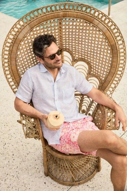 Adult man in a woven chair by a pool, wearing sunglasses and a patterned shirt, holding a coconut drink in a tropical setting.