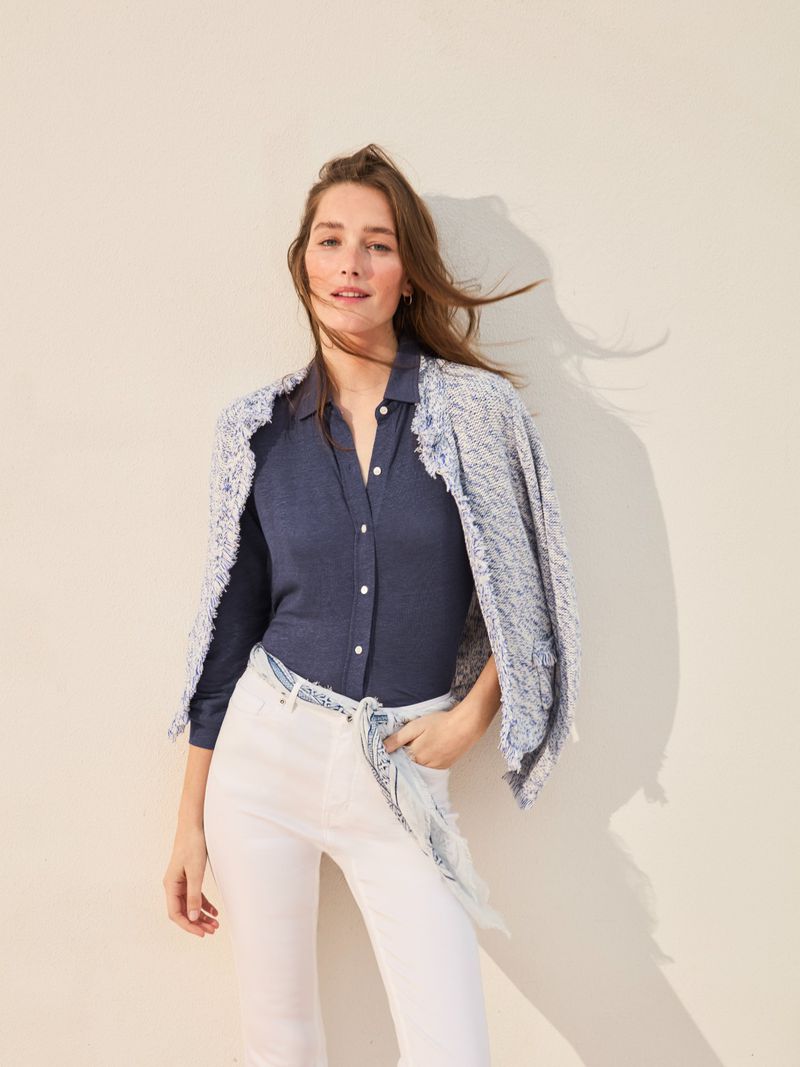Smiling woman in a blue shirt and white jeans with a jacket over her shoulders in front of a neutral background in a studio.
