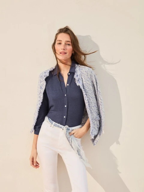 Smiling woman in a blue shirt and white jeans with a jacket over her shoulders in front of a neutral background in a studio.