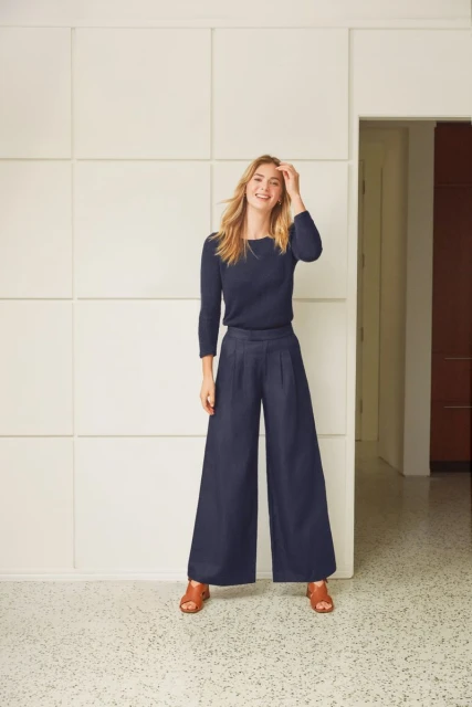 Blonde woman poses in navy sweater and wide-leg pants in front of a geometric white wall, smiling and looking at the camera.
