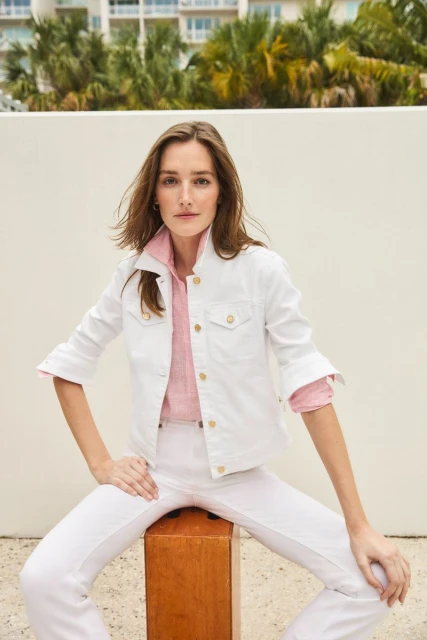 A woman in a white denim jacket and pants is seated on a wooden cube in front of a pale wall with palm trees and a building behind.