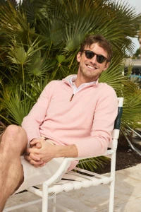 Smiling man wearing dark sunglasses, a light pink quarter-zip sweater, and white shorts seated in a white chair before palm trees.