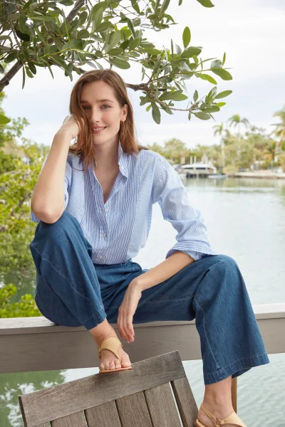 Woman in blue striped shirt and wide-leg jeans seated outdoors near water for J. McLaughlin March '24 Catalogue