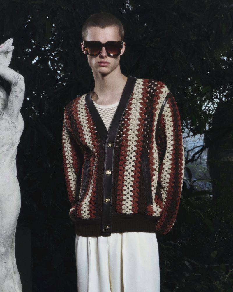 A stylish adult male wears a crochet cardigan and white pants, accessorized with dark sunglasses, posing in a dimly lit outdoor setting.