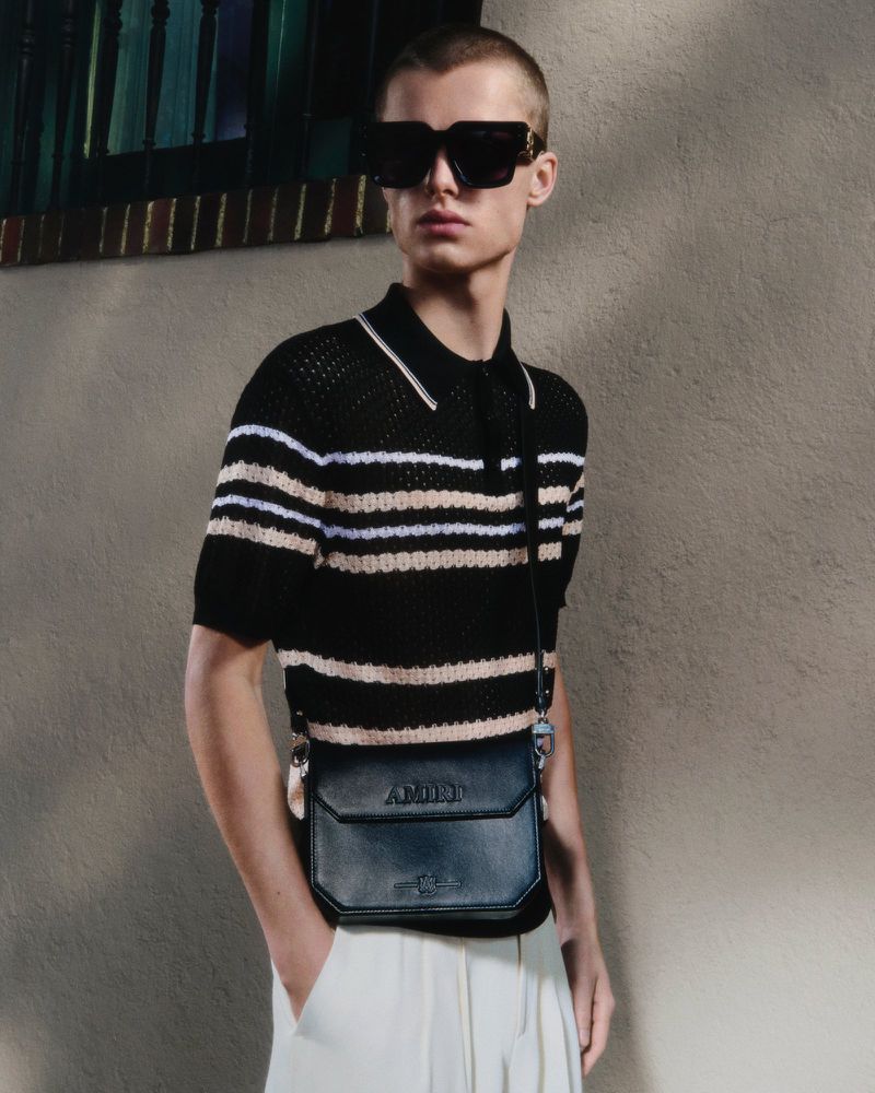 Male fashion model poses in a striped polo shirt with a small crossbody bag against a neutral wall in a studio setting.