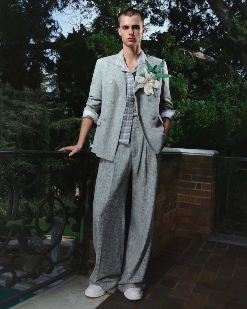 A young adult male poses in a light gray suit, standing on a brick patio with green trees in the background on a sunny day.