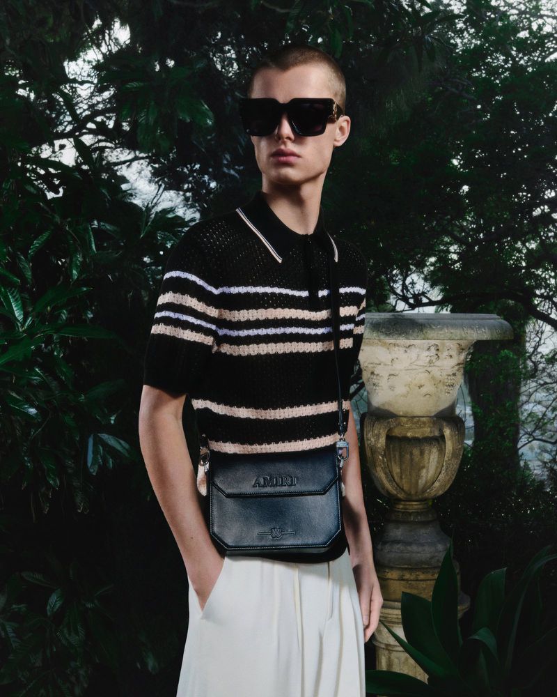 A young adult man wears sunglasses and a striped shirt in a natural setting, carrying a small black bag, showcasing outdoor fashion.