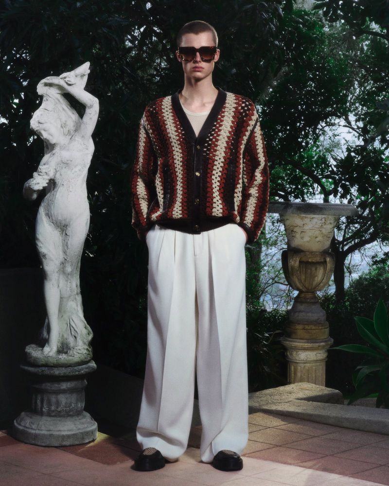 A fair-skinned male model in a striped cardigan and wide-leg trousers stands in a garden setting with statues, conveying relaxed style.