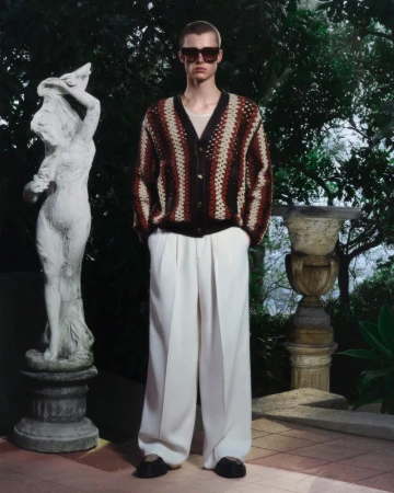 A fair-skinned male model in a striped cardigan and wide-leg trousers stands in a garden setting with statues, conveying relaxed style.