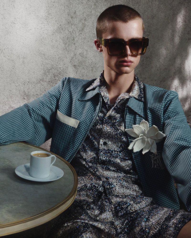 Man in a patterned shirt and blue jacket sitting at a table with an espresso. The image is backlit, creating strong shadows on the textured wall.