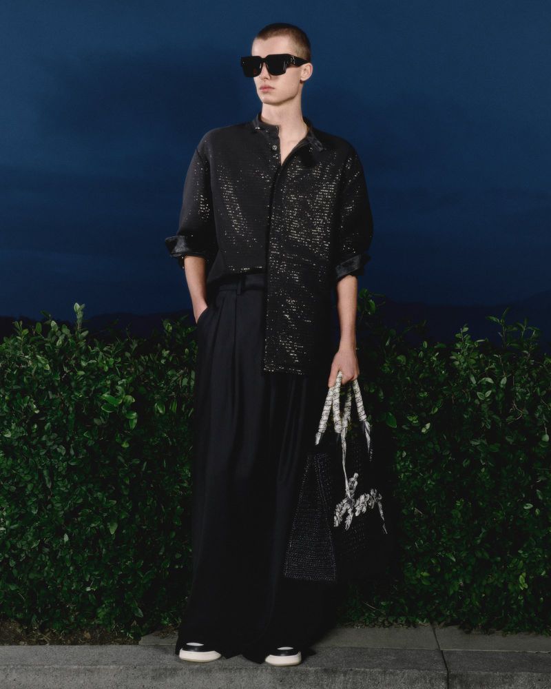 Adult man in stylish black shirt, wide-leg trousers, and sunglasses poses with a patterned tote bag against an outdoor backdrop.