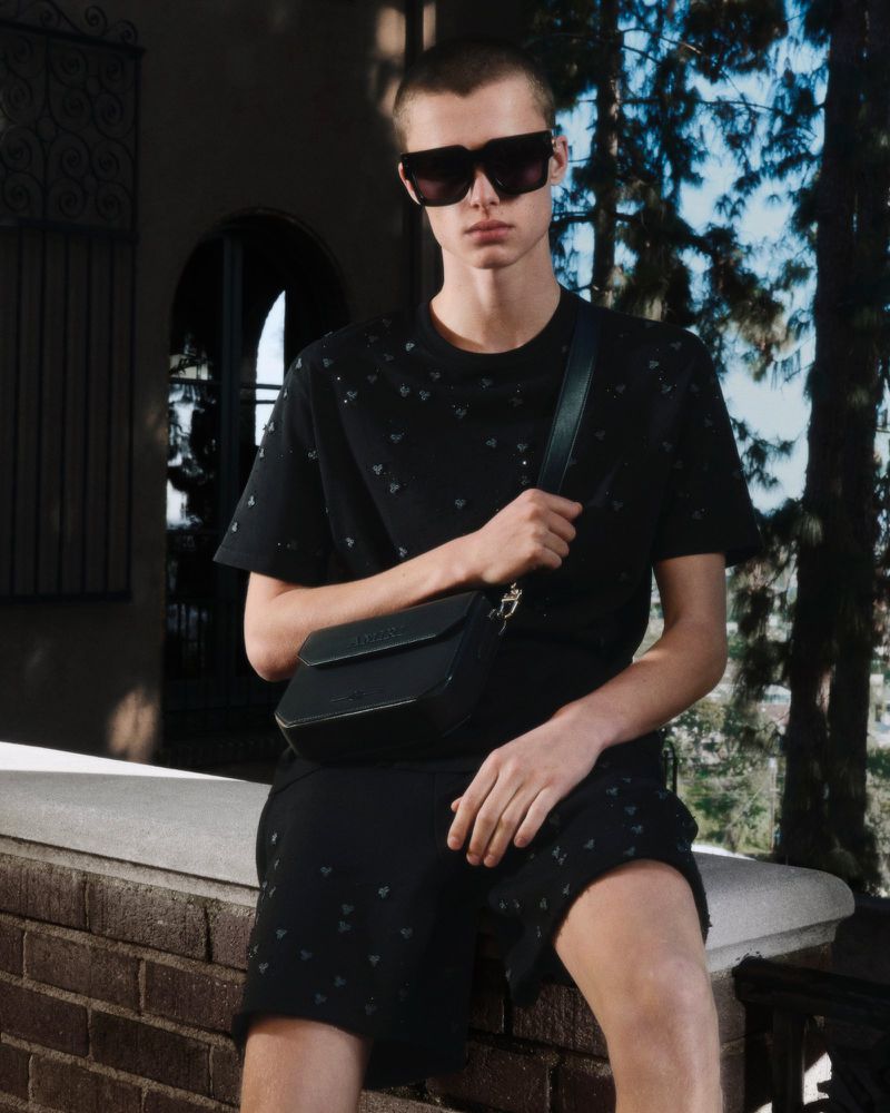 A young man wearing sunglasses, a patterned black shirt and shorts, and a shoulder bag sits on a brick wall outdoors in soft daylight.