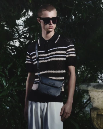 Model wearing dark sunglasses, striped polo shirt, and a crossbody bag outdoors in front of dark foliage