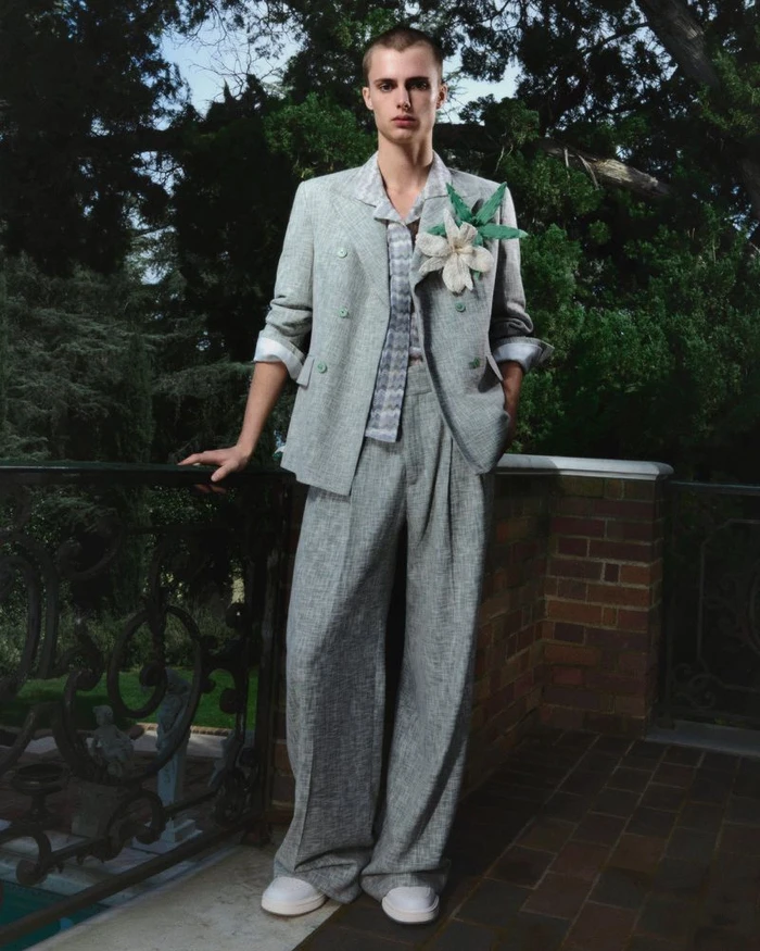 Model standing on a balcony wearing a grey double-breasted suit with a large flower lapel pin against a lush tree background.