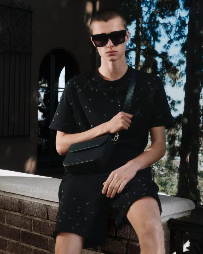 Model wearing large black sunglasses, an embellished black t-shirt, shorts, and a crossbody bag seated on a brick ledge outdoors.