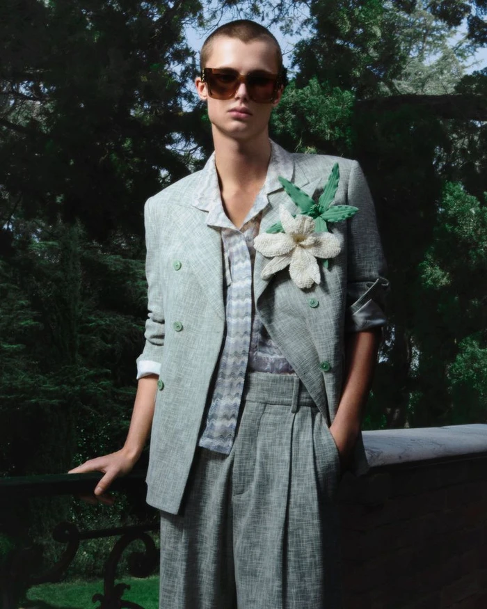 Model wearing a grey textured blazer with a large white flower lapel pin outdoors against dark trees