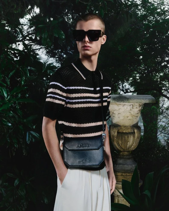 Model wearing black sunglasses, striped polo shirt, and white trousers holding a black crossbody bag outdoors near an urn.
