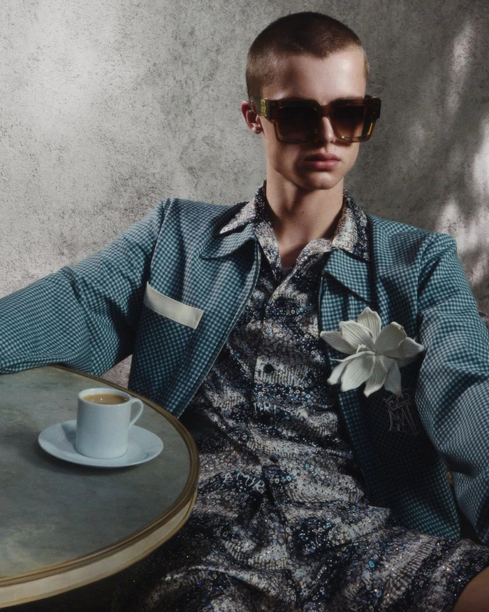 Model Elias Monstrey seated at a table wearing an Amiri teal patterned jacket, floral brooch, and dark square sunglasses.