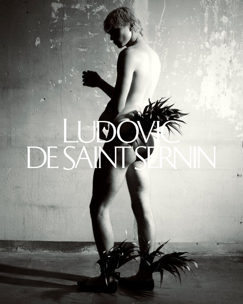 Monochrome image of a shirtless person adorned with plant leaves posing against a weathered wall with visible text overlay. 