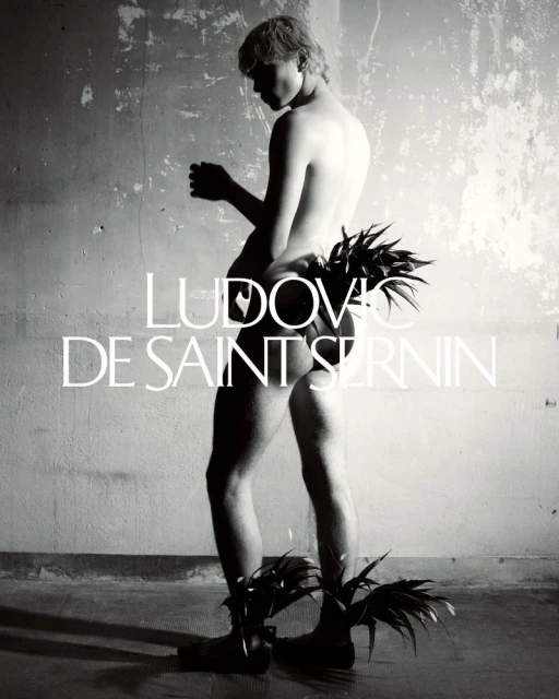 Monochrome image of a shirtless person adorned with plant leaves posing against a weathered wall with visible text overlay. 