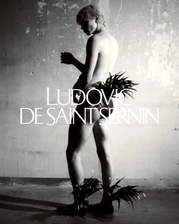 Monochrome image of a shirtless person adorned with plant leaves posing against a weathered wall with visible text overlay. 