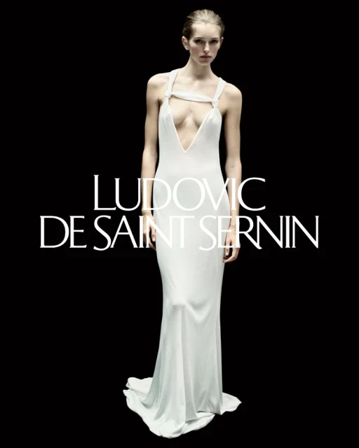 Fashion shot of a woman modeling a white gown with a plunging neckline against a black background. Text reads: LUDOVIC DE SAINT SERNIN.