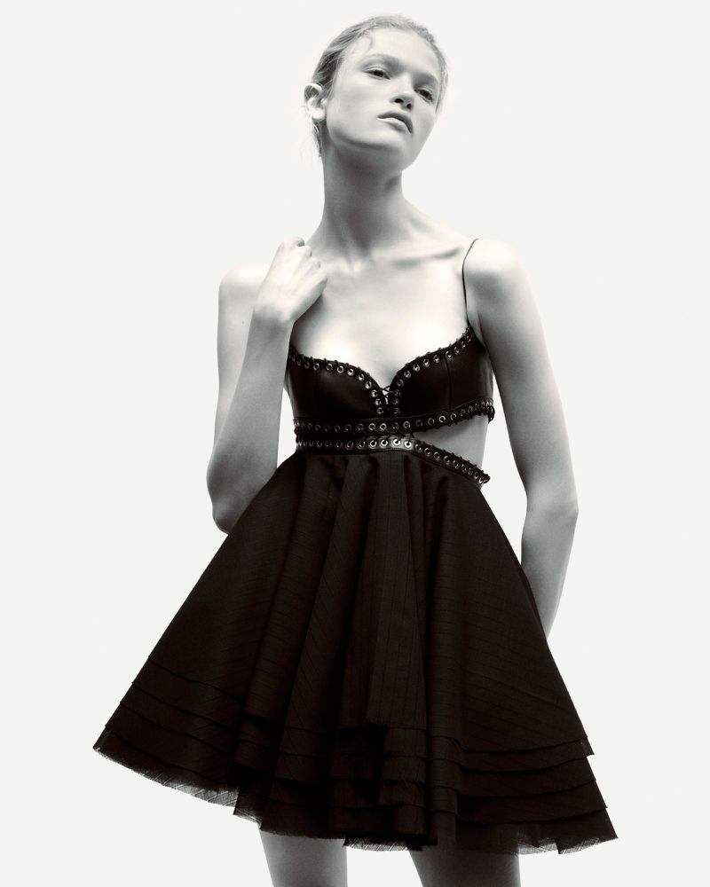 Fashion photo of a slender woman in a short black dress with a cut-out bodice, posing in front of a white background with soft, diffused lighting.