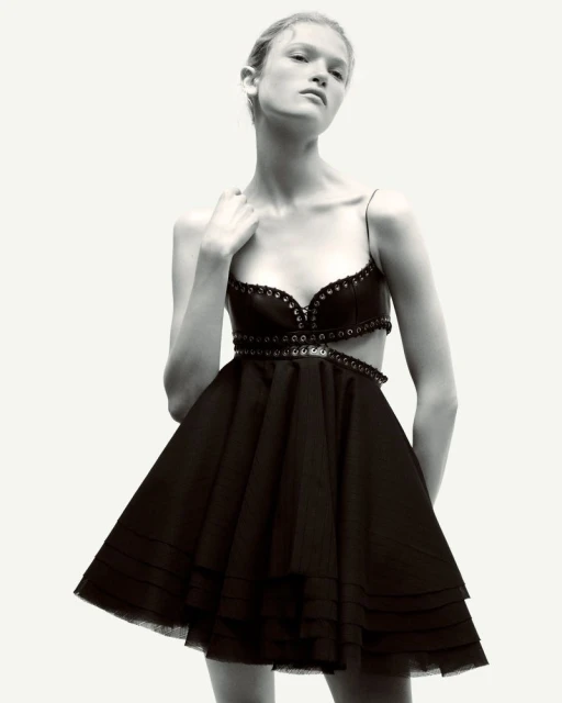 Fashion photo of a slender woman in a short black dress with a cut-out bodice, posing in front of a white background with soft, diffused lighting.