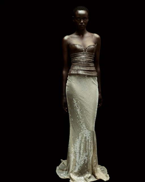 A solitary Black female fashion model poses in a strapless, beaded top and shimmering champagne-colored gown against a stark black background.