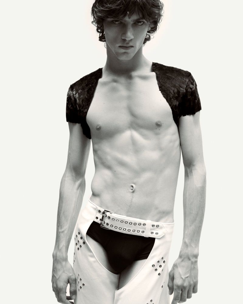 Male fashion model poses shirtless in white chaps with dark briefs against a neutral backdrop in a studio shot with moody lighting.